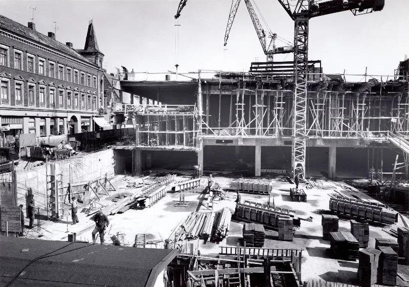 A black-and-white photo of a building site in a city, with a crane and building materials. 