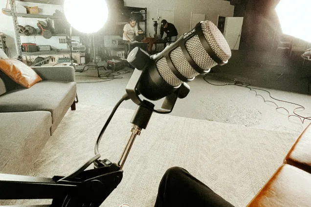 A photo of a microphone. Behind it a room with lights, sofas, and two people working at a desk.