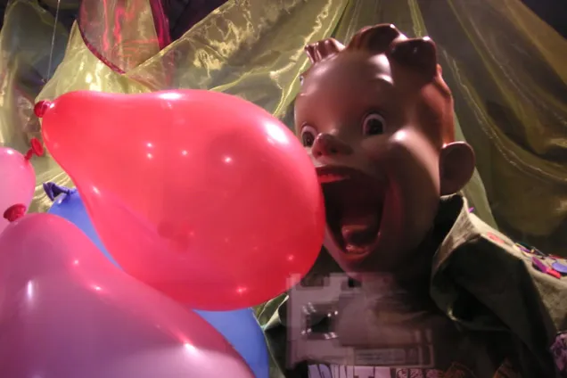 A photo of a colourful plastic doll, gaping at a red ballon.
