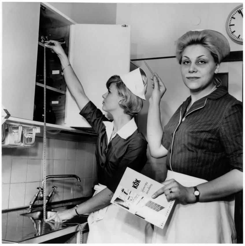 Svartvit bild på två personer i ett kök. Personerna har uniform med förkläde och hätta.