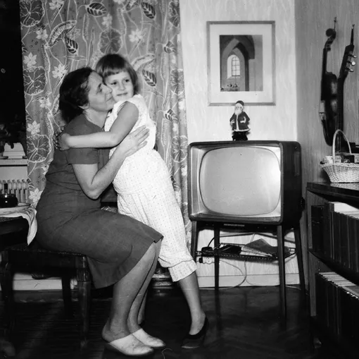 Svartvitt fotografi av två personer bredvid en tv-apparat. En vuxen personen sitter ner, och kramar ett barn som står upp.