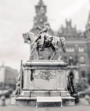 Fotografi av en staty. Statyn har en stor sockel med inskription. Ovanpå sockeln är en häst med ryttare.